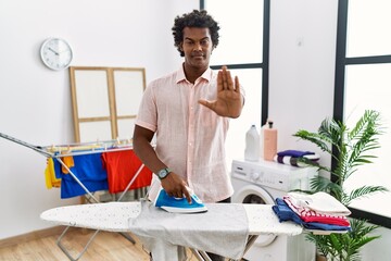 African man with curly hair ironing clothes at home doing stop sing with palm of the hand. warning expression with negative and serious gesture on the face.