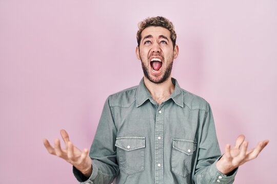 Hispanic man with beard standing over pink background crazy and mad shouting and yelling with aggressive expression and arms raised. frustration concept.