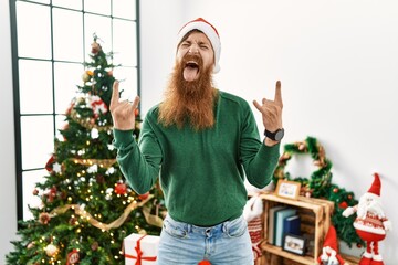 Redhead man with long beard wearing christmas hat by christmas tree shouting with crazy expression...