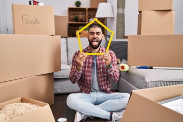 Middle east man with beard sitting on the floor at new home holding ruler sticking tongue out happy...