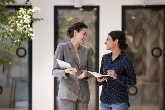 Two attractive multi ethnic businesswomen enjoy success, sales growth and business advancement smile enjoy conversation and career in company standing in office hallway, Friendship at work, workflow