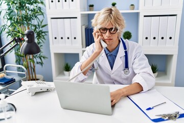 Young blond man doctor using laptop talking on telephone at clinic