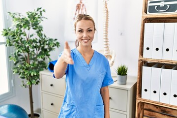 Young caucasian woman working at pain recovery clinic smiling friendly offering handshake as greeting and welcoming. successful business.