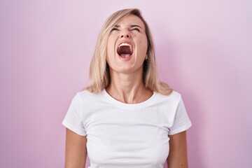 Young blonde woman standing over pink background angry and mad screaming frustrated and furious, shouting with anger. rage and aggressive concept.