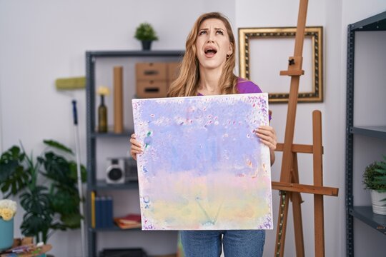 Beautiful blonde woman holding painting canvas angry and mad screaming frustrated and furious, shouting with anger looking up.