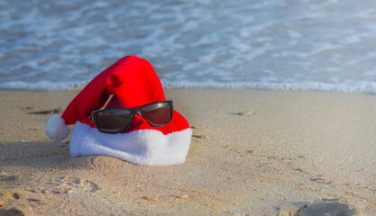 Christmas and New Year on the beach by the sea while relaxing and traveling to warm countries. Red Santa Claus hat with sunglasses on the sand by the sea.