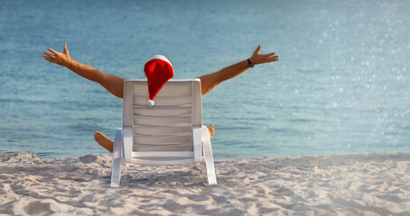 A man in a Santa Claus hat is sitting on a chaise longue by the sea, joyfully spreading his arms...