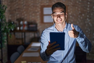 Handsome hispanic man working at the office at night doing happy thumbs up gesture with hand. approving expression looking at the camera showing success.