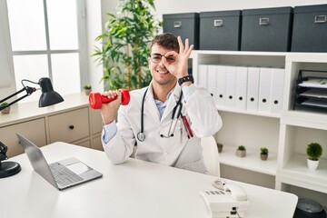 Young hispanic doctor man holding dumbbells smiling happy doing ok sign with hand on eye looking through fingers