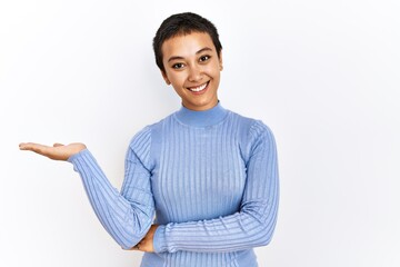 Young hispanic woman with short hair standing over isolated background smiling cheerful presenting and pointing with palm of hand looking at the camera.