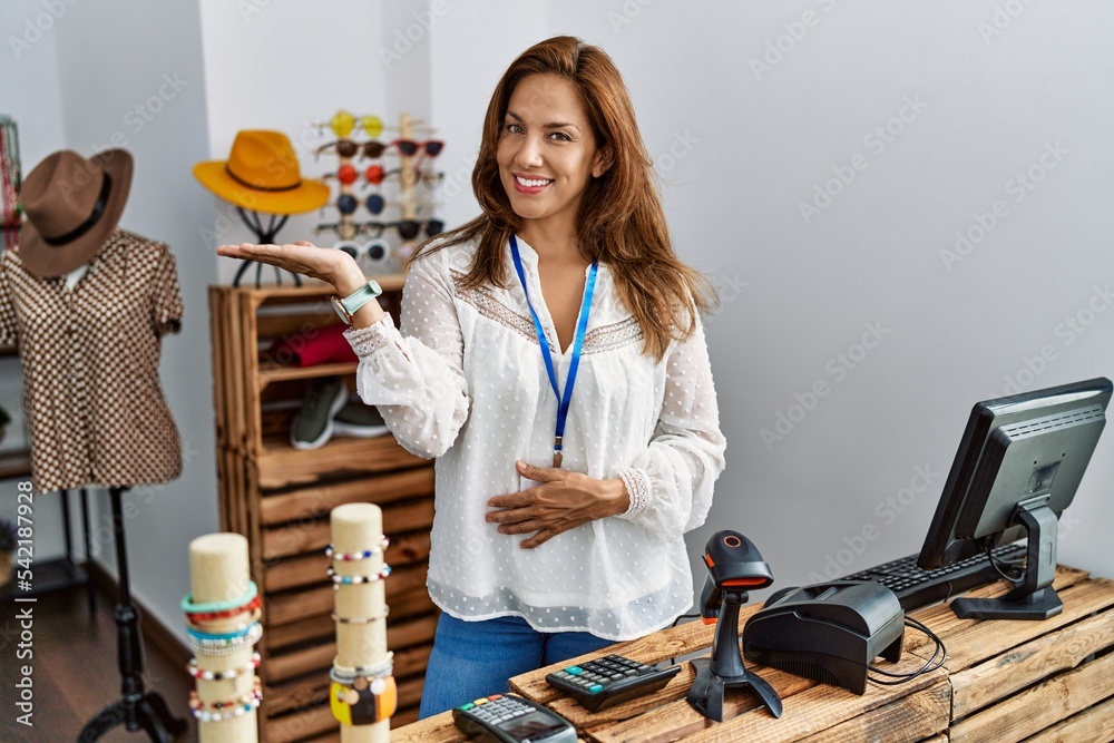 Sticker Middle age hispanic woman working as manager at retail boutique smiling cheerful presenting and pointing with palm of hand looking at the camera.