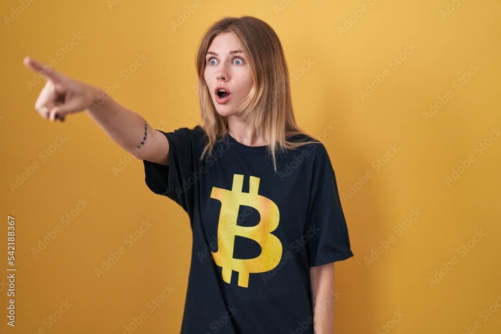 Poster Blonde caucasian woman wearing bitcoin t shirt pointing with finger surprised ahead, open mouth amazed expression, something on the front