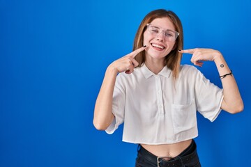 Beautiful woman standing over blue background smiling cheerful showing and pointing with fingers teeth and mouth. dental health concept.