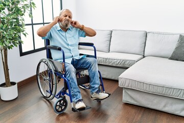 Handsome senior man sitting on wheelchair at the living room covering ears with fingers with...