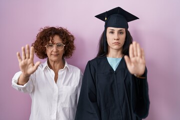 Hispanic mother and daughter wearing graduation cap and ceremony robe doing stop sing with palm of the hand. warning expression with negative and serious gesture on the face.