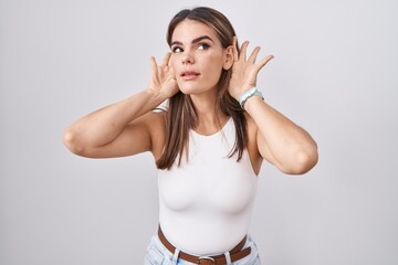 Obraz na płótnie Canvas Hispanic young woman standing over white background trying to hear both hands on ear gesture, curious for gossip. hearing problem, deaf