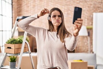 Beautiful woman holding keys of new home showing on video call puffing cheeks with funny face. mouth inflated with air, catching air. - obrazy, fototapety, plakaty