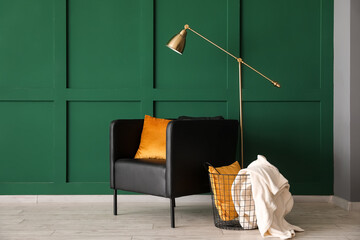 Black armchair, basket with cushion, plaid and lamp near green wall