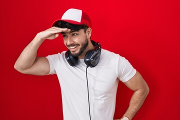 Fototapeta premium Hispanic man with beard wearing gamer hat and headphones very happy and smiling looking far away with hand over head. searching concept.