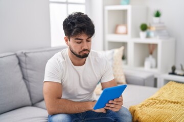 Hispanic man with beard using touchpad sitting on the sofa depressed and worry for distress, crying angry and afraid. sad expression.