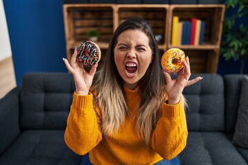 Young hispanic woman eating doughnuts at home angry and mad screaming frustrated and furious,...