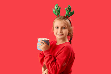 Happy little girl in reindeer horns with cup of cocoa on red background