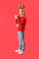 Happy little girl in reindeer horns with cup of cocoa on red background