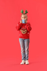 Happy little girl in reindeer horns drinking cocoa on red background