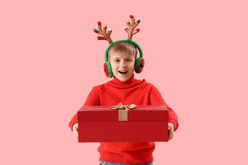 Happy little boy in reindeer horns with Christmas gift on pink background