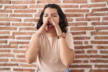 Young hispanic woman standing over bricks wall shouting angry out loud with hands over mouth