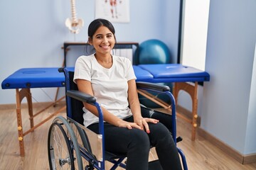 Young hispanic woman sitting on wheelchair at physiotherapy clinic with a happy and cool smile on...
