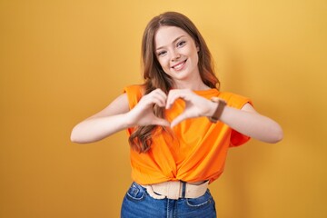 Caucasian woman standing over yellow background smiling in love doing heart symbol shape with hands. romantic concept.
