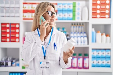 Young blonde woman pharmacist holding pills bottle talking on smartphone at pharmacy