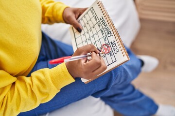 African american woman writing on calendar sitting on bed at bedroom