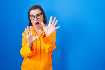 Middle age hispanic woman wearing glasses standing over blue background afraid and terrified with fear expression stop gesture with hands, shouting in shock. panic concept.