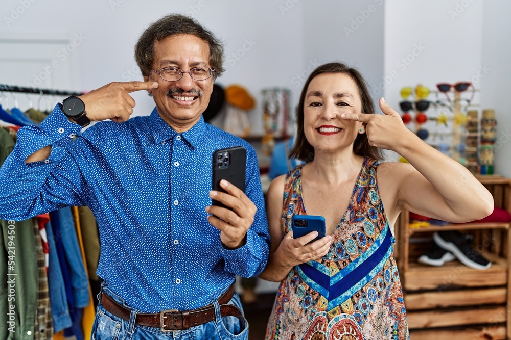 Sticker Middle age interracial couple at retail shop using smartphone pointing with hand finger to face and nose, smiling cheerful. beauty concept