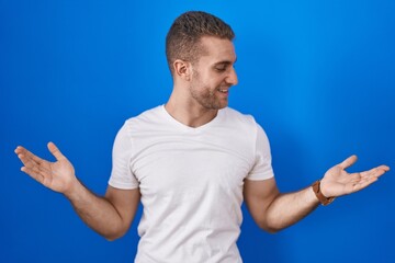 Young caucasian man standing over blue background smiling showing both hands open palms, presenting...
