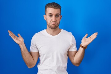 Fototapeta premium Young caucasian man standing over blue background clueless and confused expression with arms and hands raised. doubt concept.