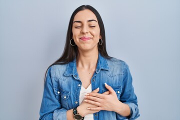 Hispanic woman standing over blue background smiling with hands on chest with closed eyes and...