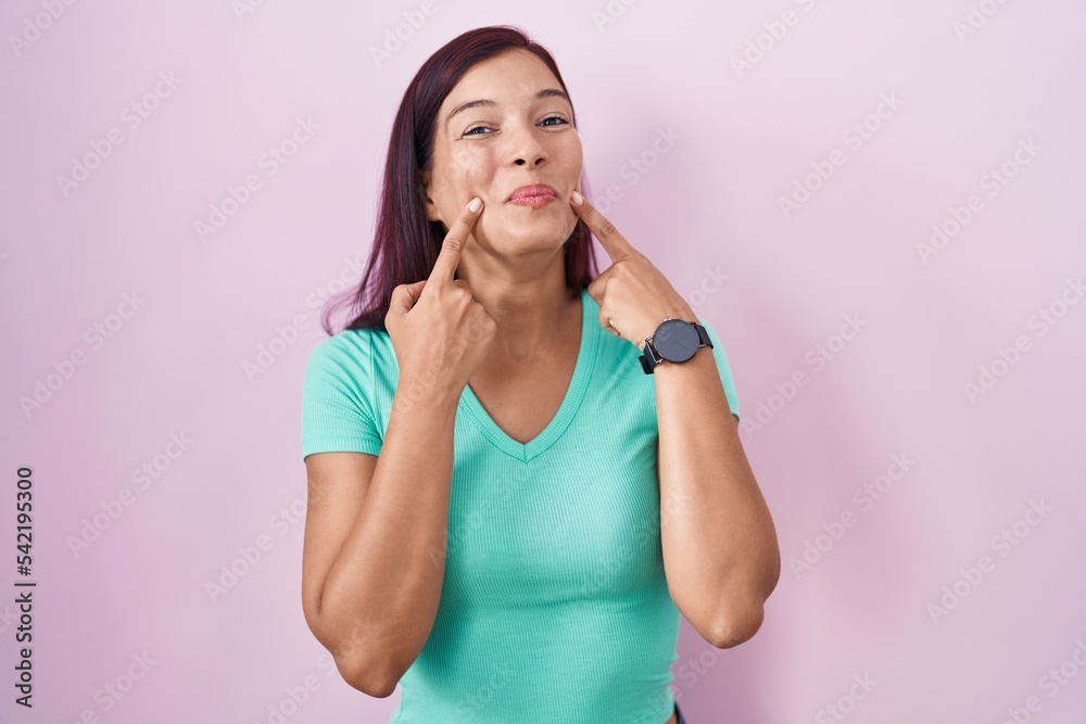 Sticker Young hispanic woman standing over pink background smiling with open mouth, fingers pointing and forcing cheerful smile