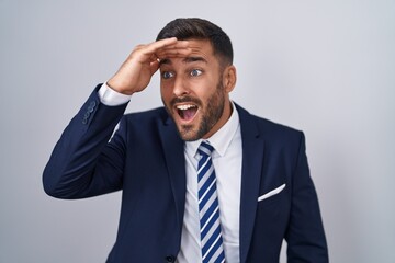 Handsome hispanic man wearing suit and tie very happy and smiling looking far away with hand over head. searching concept.