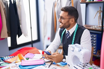 Young hispanic man tailor smiling confident using smartphone at tailor shop