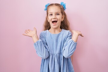 Young little girl standing over pink background celebrating victory with happy smile and winner expression with raised hands