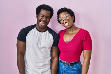 Obraz premium Young african american couple standing over pink background sticking tongue out happy with funny expression. emotion concept.