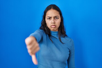 Obraz premium Young brazilian woman standing over blue isolated background looking unhappy and angry showing rejection and negative with thumbs down gesture. bad expression.