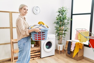 Young caucasian woman smiling confident holding basket with clothes at laundry room
