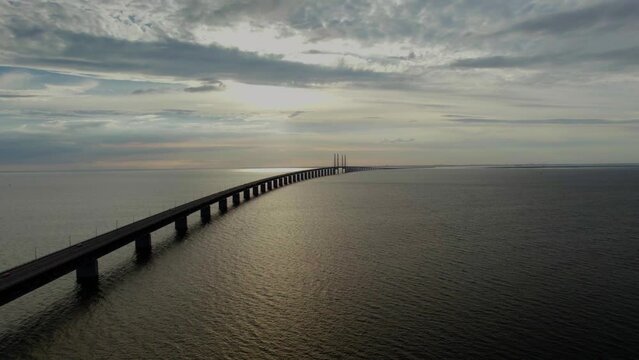 High angle aerial arc shot of the Öresundsbron bridge between Sweden and Denmark over the Öresund strait near Malmö. Colorful sunset reflected on the calm waves, with cars driving on the bridge