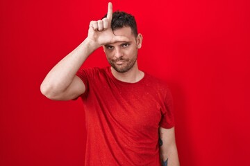 Young hispanic man standing over red background making fun of people with fingers on forehead doing...