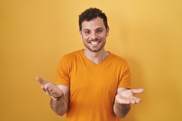 Young hispanic man standing over yellow background smiling cheerful offering hands giving assistance and acceptance.
