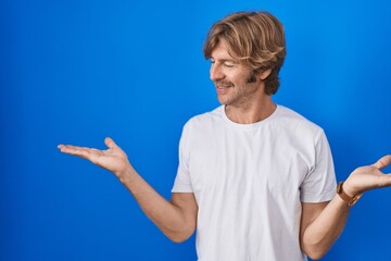 Middle age man standing over blue background smiling showing both hands open palms, presenting and...